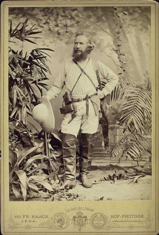 Vintage studio portrait of a bearded man in a tropical setting, wearing a hat and carrying a gun.
