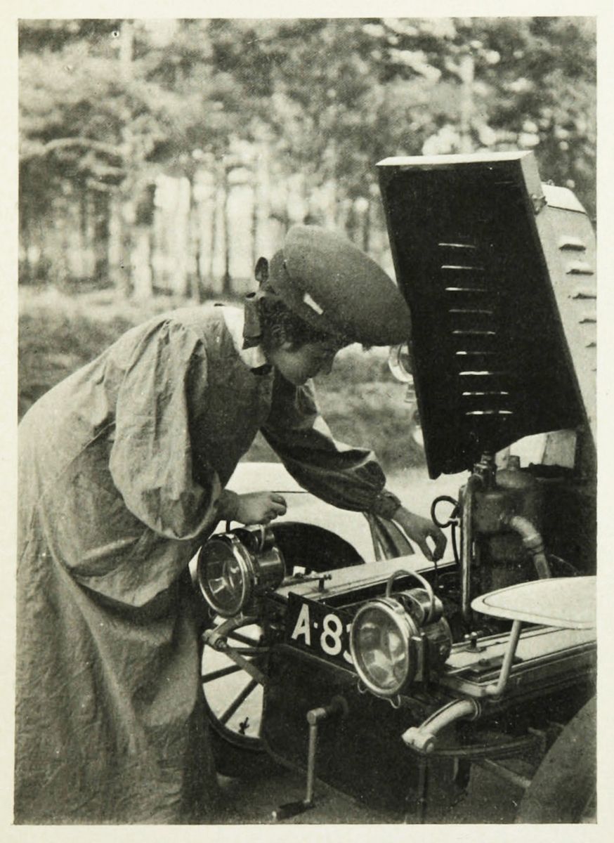 A vintage black and white photograph of a person wearing a cap and goggles working on the engine of a classic car in a wooded area.