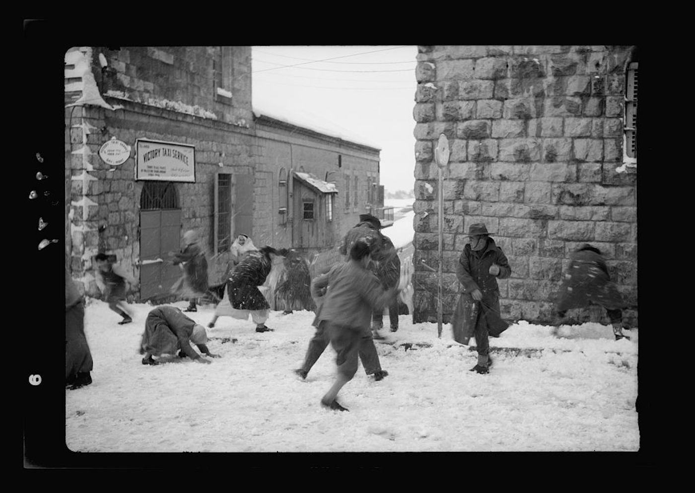 A black and white photograph of a snowball fight in a snowy urban setting.