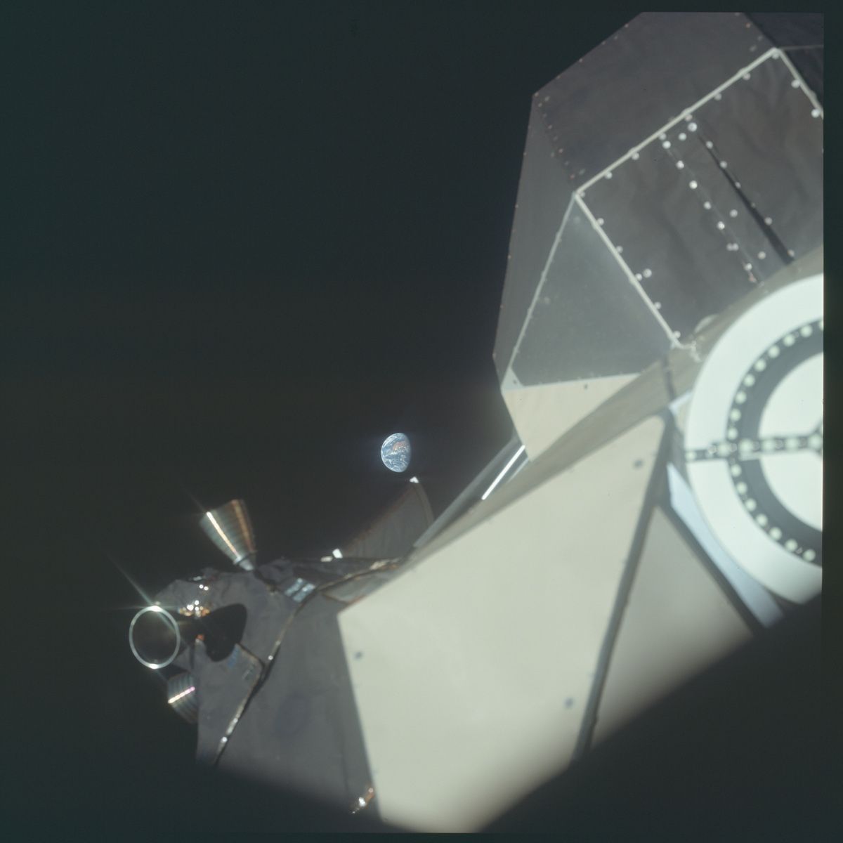 A view from a spacecraft in space, looking out towards Earth. The Earth appears as a small, blue and white marble suspended in the blackness of space. The foreground is dominated by parts of the spacecraft itself, including large, flat, metallic panels and a complex arrangement of instruments and equipment.