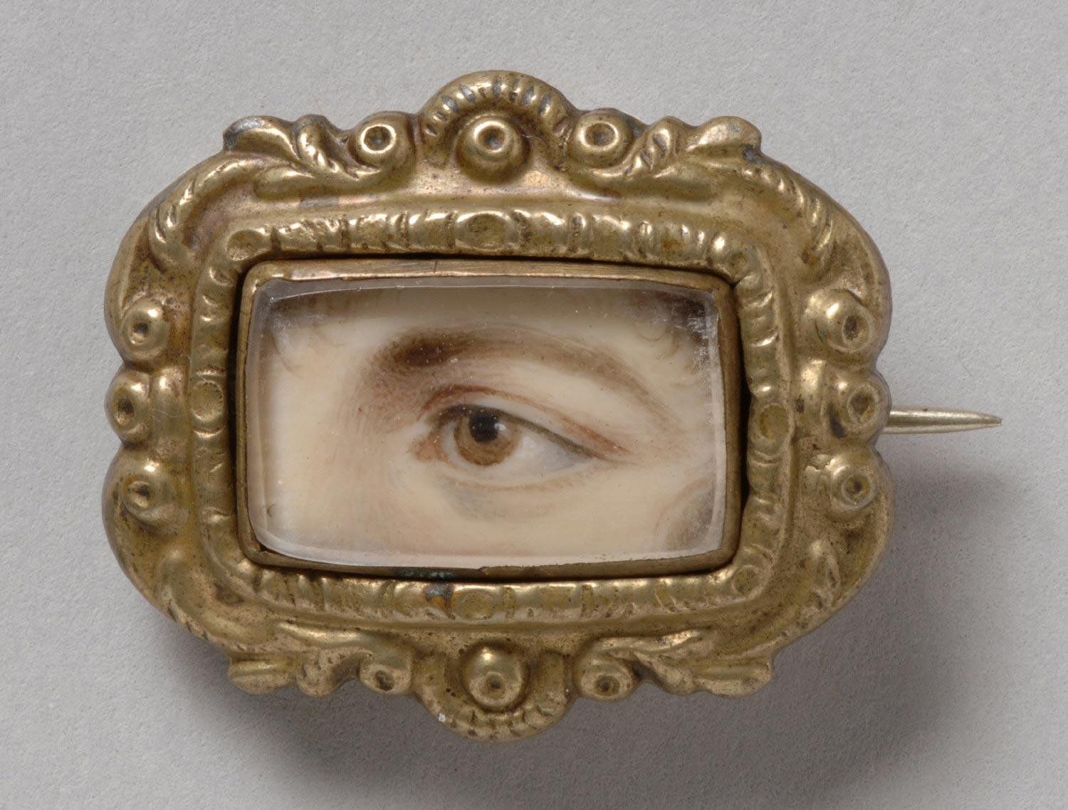 Close-up of a vintage brooch featuring a realistic depiction of a human eye set within an ornate, gilded metal frame.