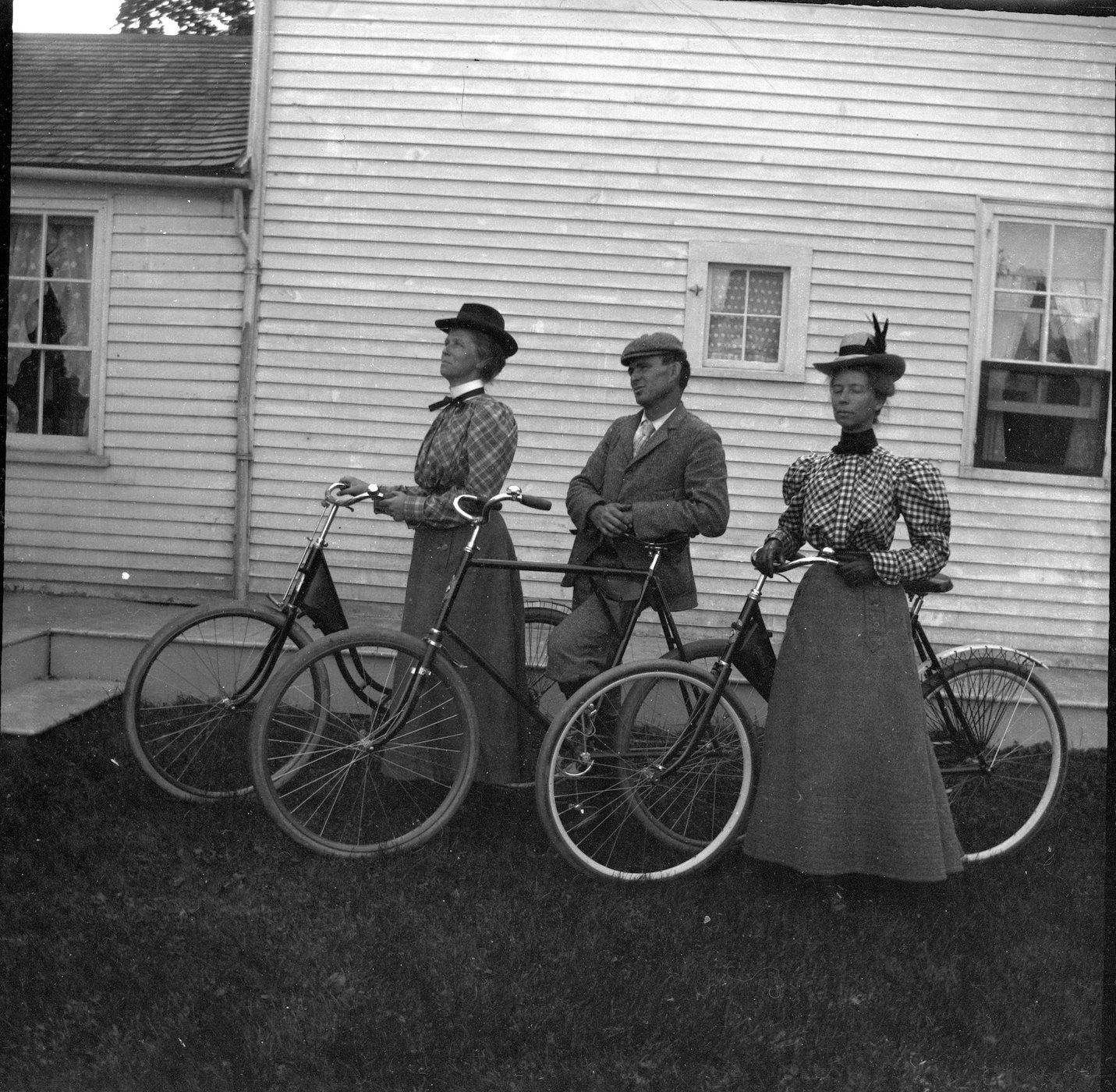 A vintage black and white photograph featuring three people with bicycles in front of a wooden-sided building.