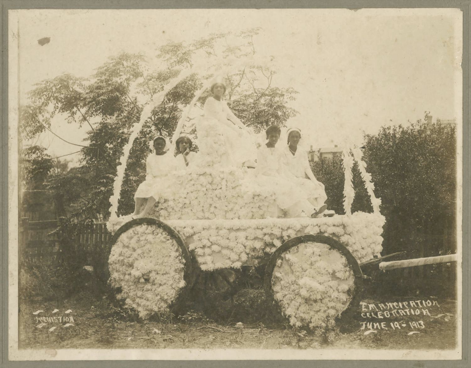 A vintage black and white photograph of an Emancipation Celebration parade float covered in white cotton, with people seated and standing on it, and a sign indicating the date as June 19, 1913.