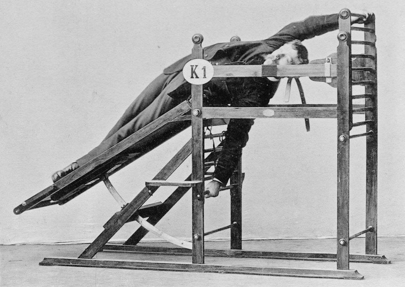A black and white photograph of a person lying on a wooden apparatus, possibly a medical device, with a sign labeled 'K1'.