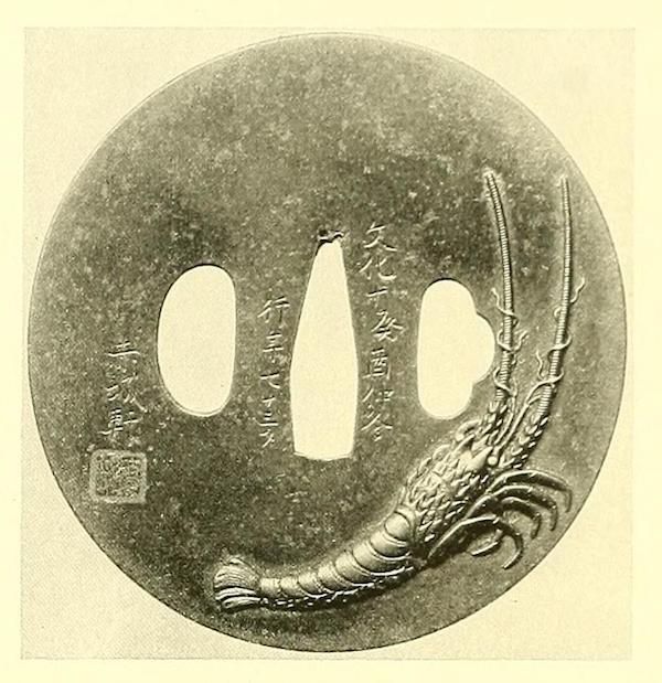 A circular tsuba with a detailed shrimp carving and Japanese text.
