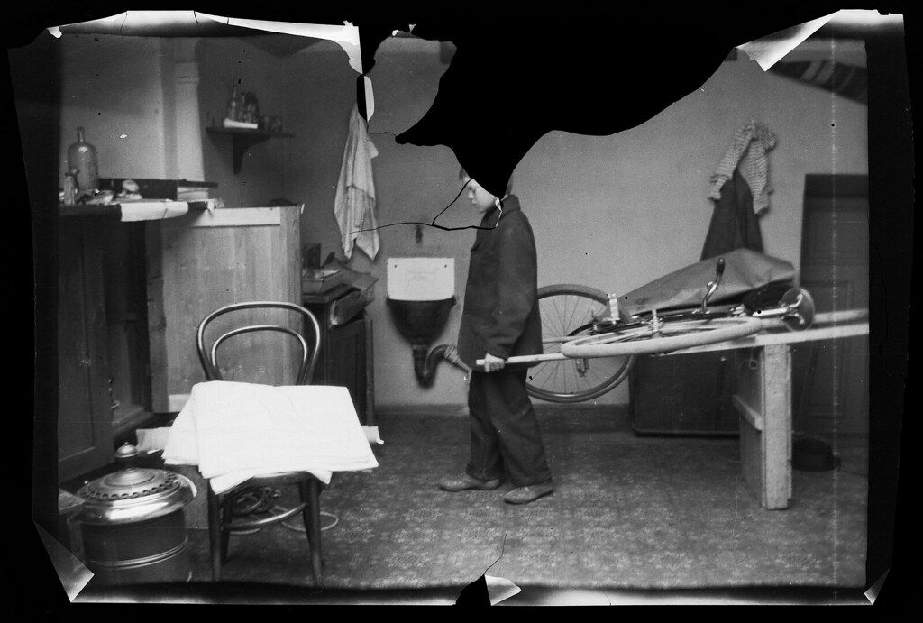 A black and white photograph of a man working on a bicycle in a cluttered workshop.