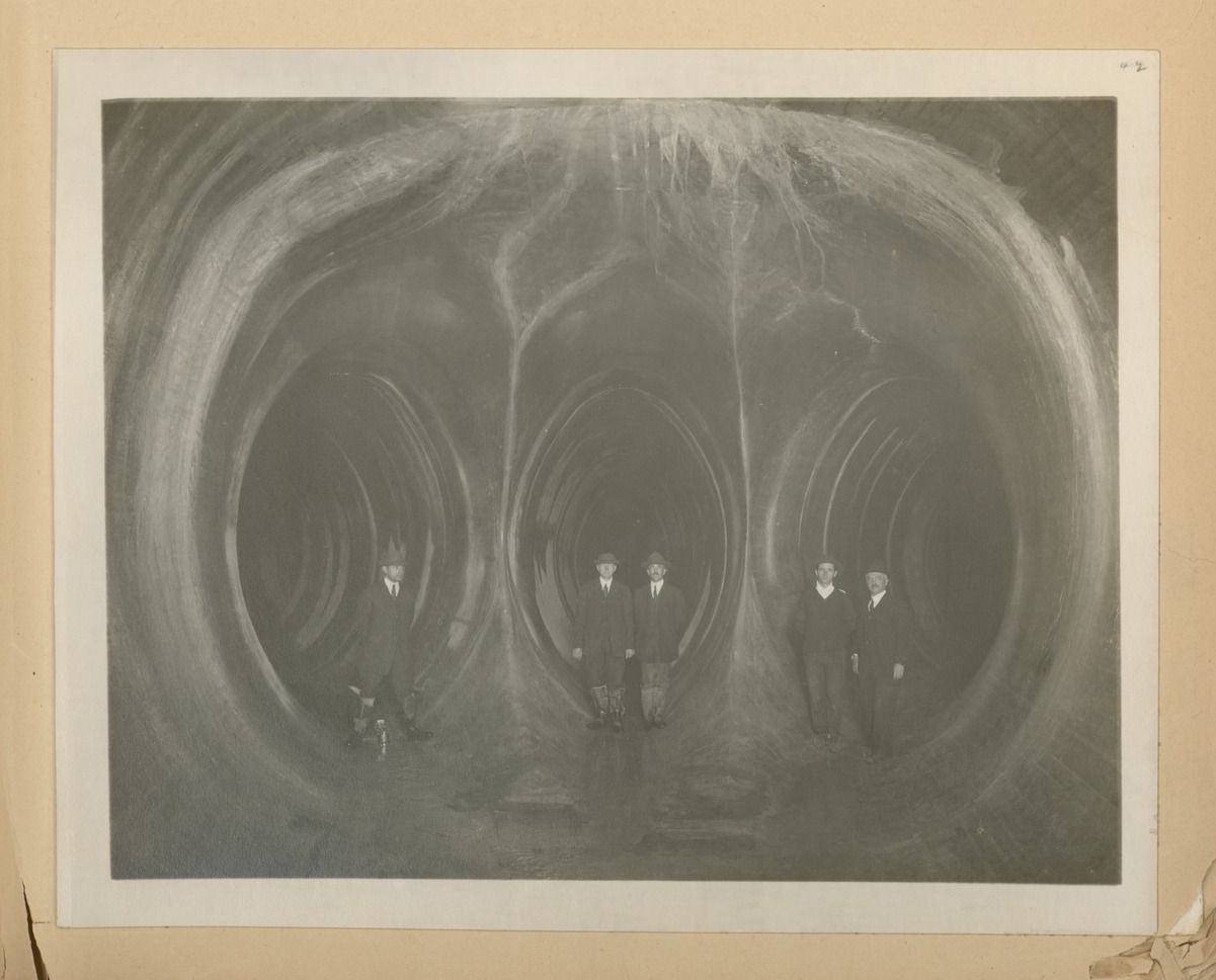 A vintage black and white photograph depicting a scientific demonstration of magnetic fields. The image shows a group of men standing in front of a dark background, with visible magnetic field lines emanating from two poles.