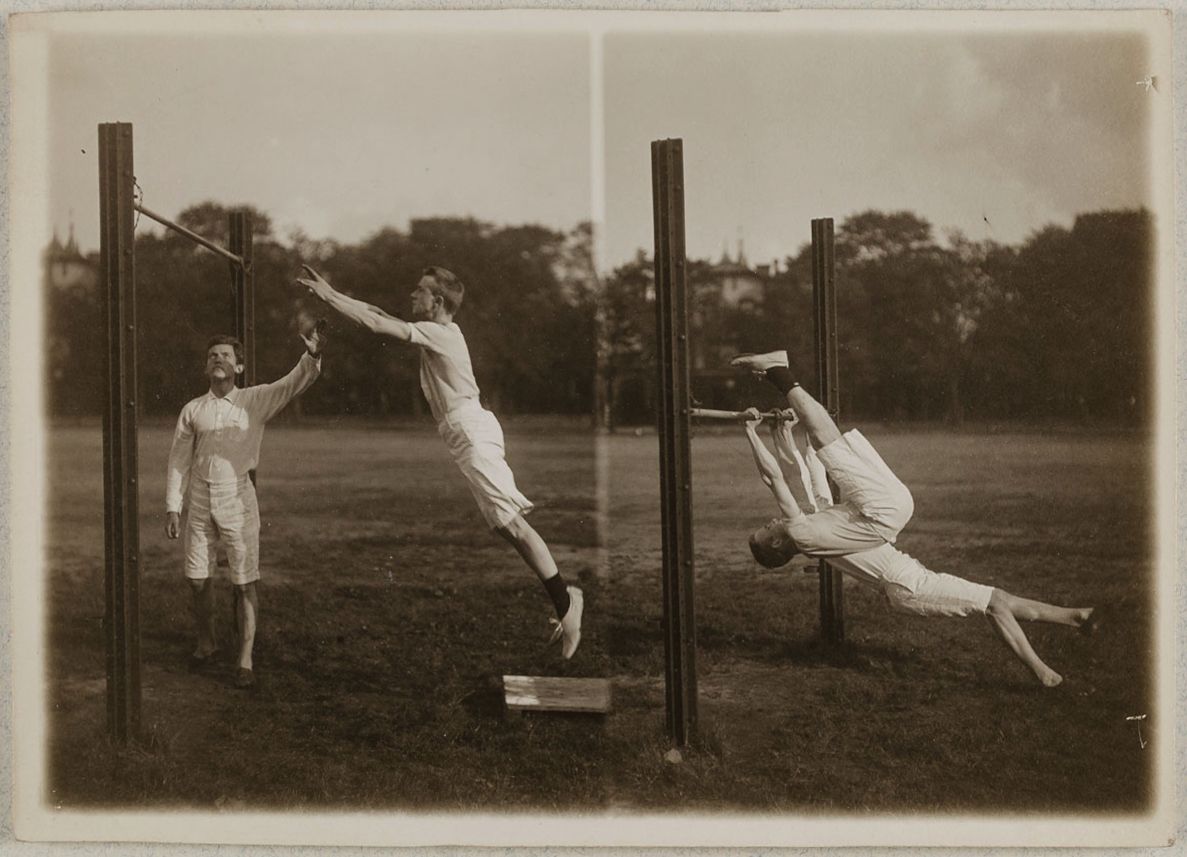 A vintage black and white photograph of three men performing a gymnastic move on parallel bars in a grassy field.