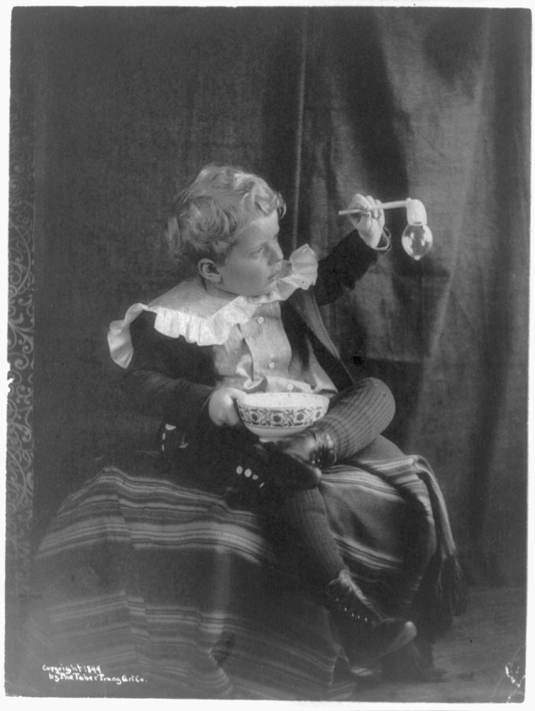 A vintage black and white photograph of a young child eating from a bowl with chopsticks.
