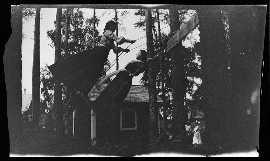 A vintage black and white photograph of children flying a large kite from the roof of a small building, surrounded by trees.