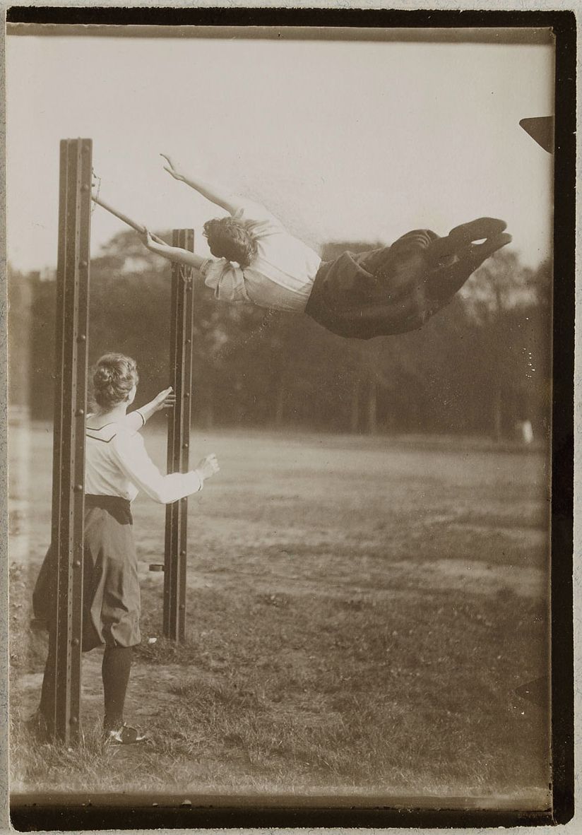 A vintage black and white photograph of a woman swinging high in the air, observed by another woman standing near a wooden swing set.