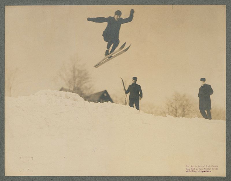 Vintage black and white photograph of a man skiing and jumping in the snow, with two other men standing nearby.