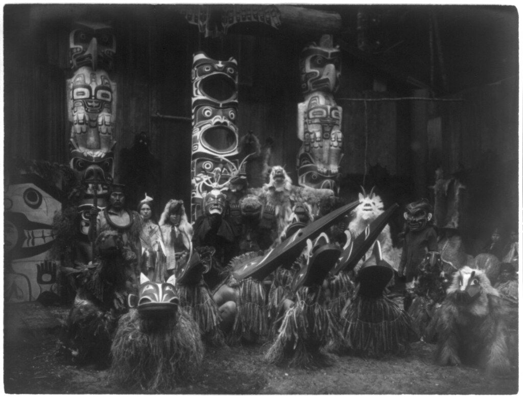 A black and white photograph of a group of people in masks and costumes performing a ceremony before a totem pole.