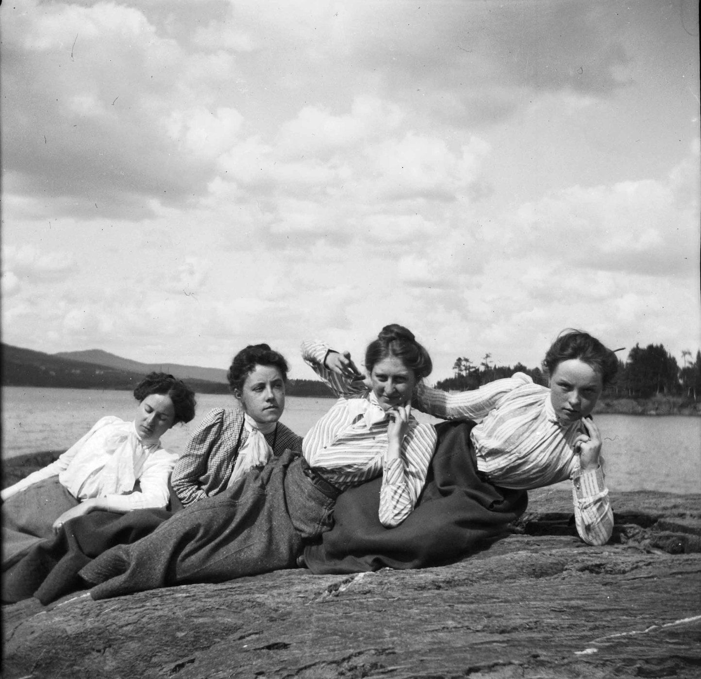 A black and white photograph of four young women lying on a rocky shoreline, looking directly at the camera.
