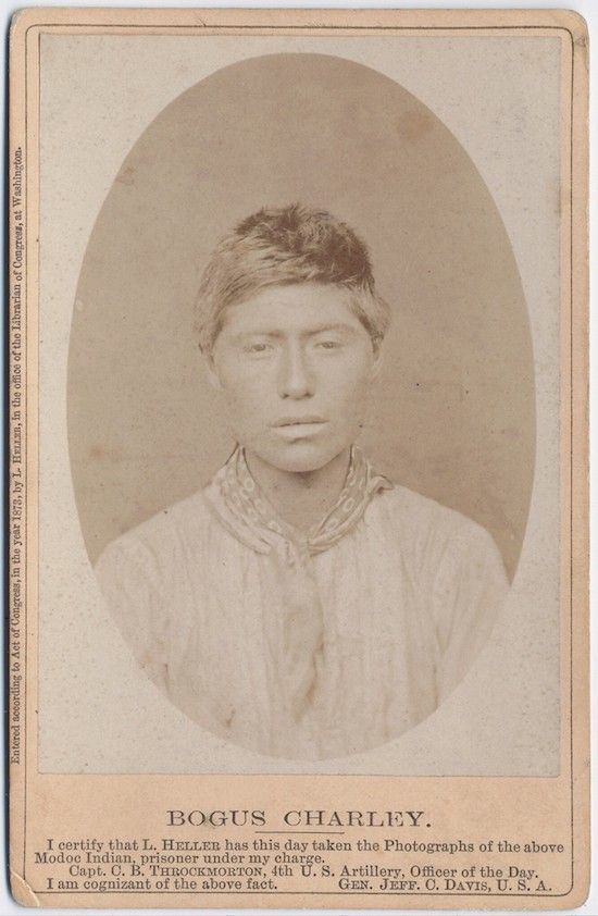 A sepia-toned photograph of a Native American man named Bogus Charley, identified as a Modoc Indian prisoner, with text below the image certifying the photograph was taken by L. Heller in 1873.