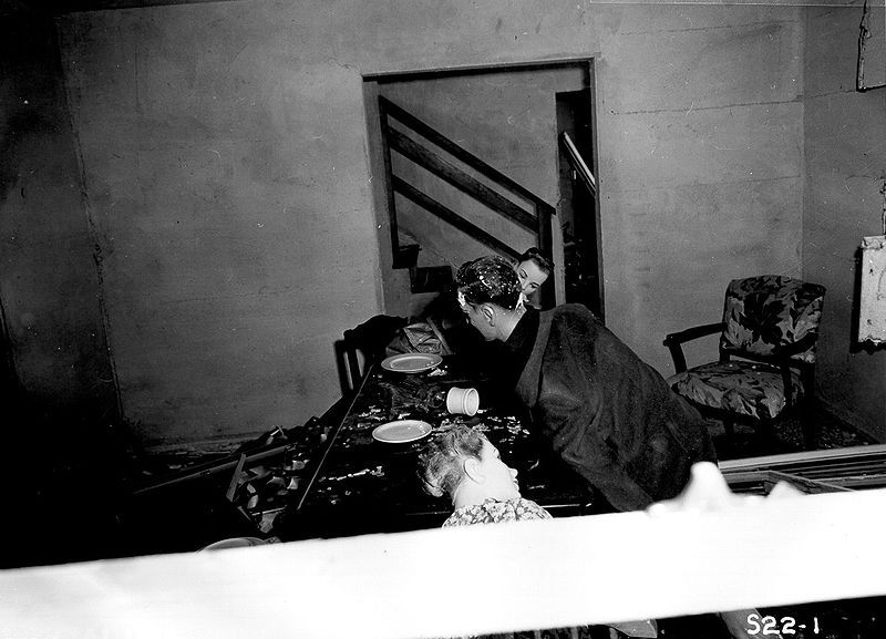 A black and white photograph of a man in a robe hunched over a table with plates and a doll in a dilapidated room.