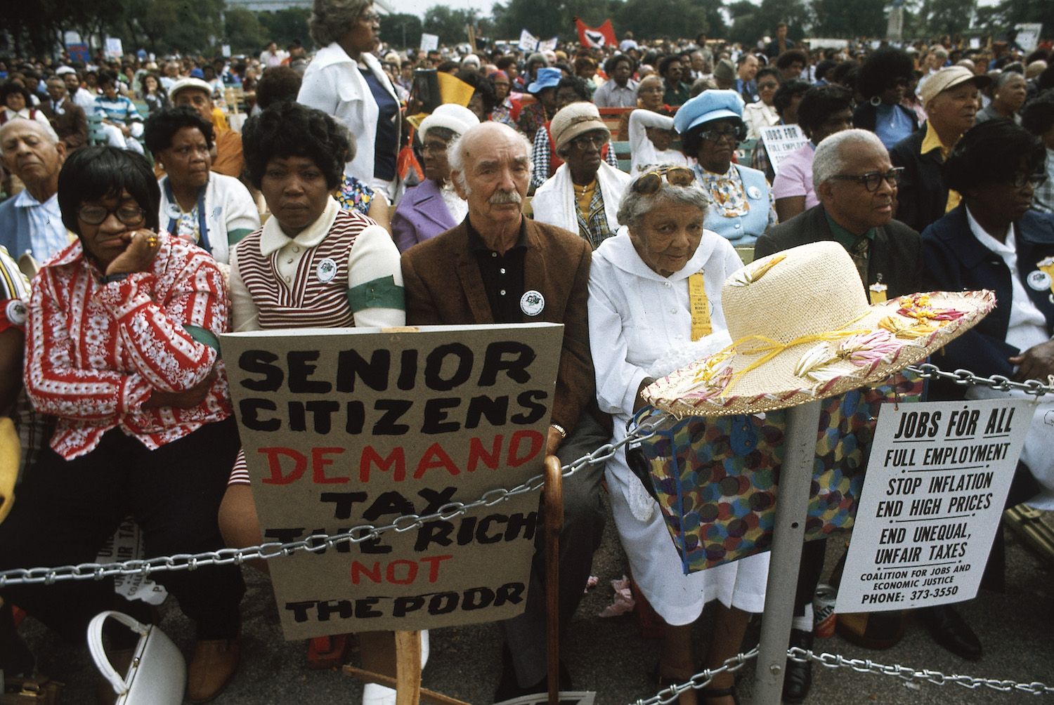A large crowd of people, mostly senior citizens, protesting with signs and banners. The signs read "Senior Citizens Demand Tax the Rich Not the Poor" and "Jobs for All, Full Employment, Stop Inflation, End High Prices, End Unequal, Unfair Taxes". The crowd is diverse and energetic, and the scene is set on a sunny street.
