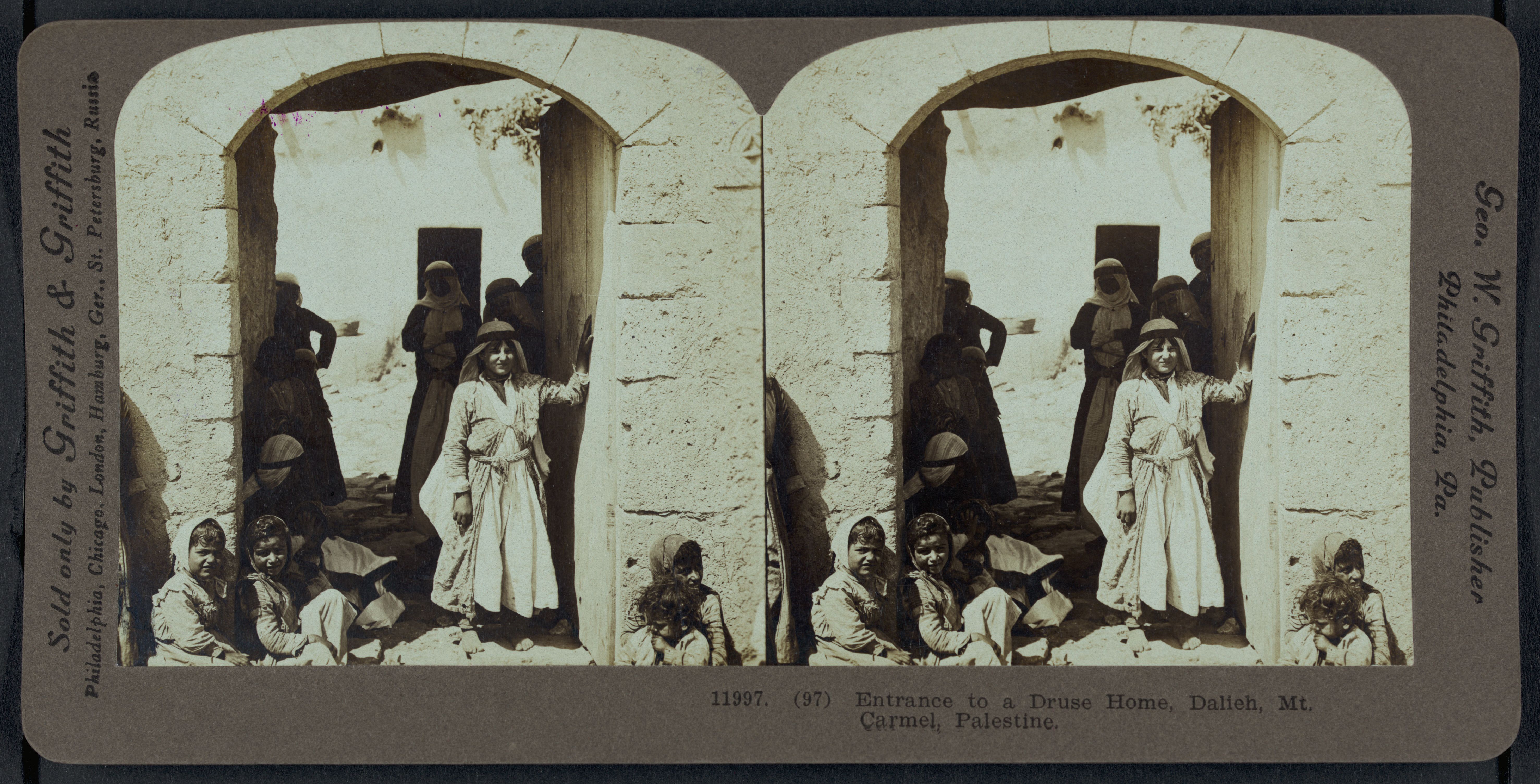 Stereoscopic photograph of an entrance to a Druse home in Dalieh, Mt. Carmel, Palestine. The image shows a stone archway leading into a building. Several people are gathered in front of the archway, some seated and some standing. They are dressed in traditional clothing, including long robes and head coverings. The landscape in the background features mountains and a rocky terrain.