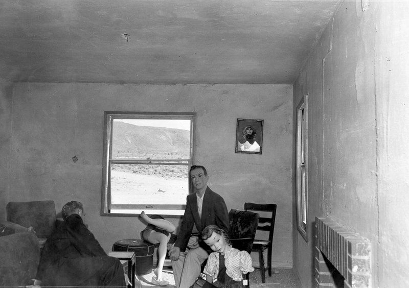 A black and white photograph of a man, a young girl, and a woman in a sparsely furnished room with a view of a desert landscape through a window.