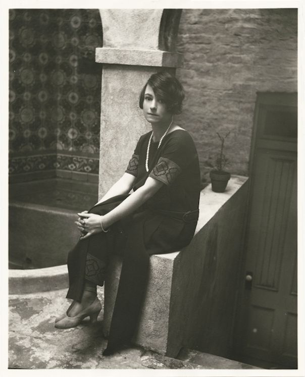 A black and white photograph of a woman seated on a stone chair, wearing a patterned dress and pearls, with a textured wall and door in the background.