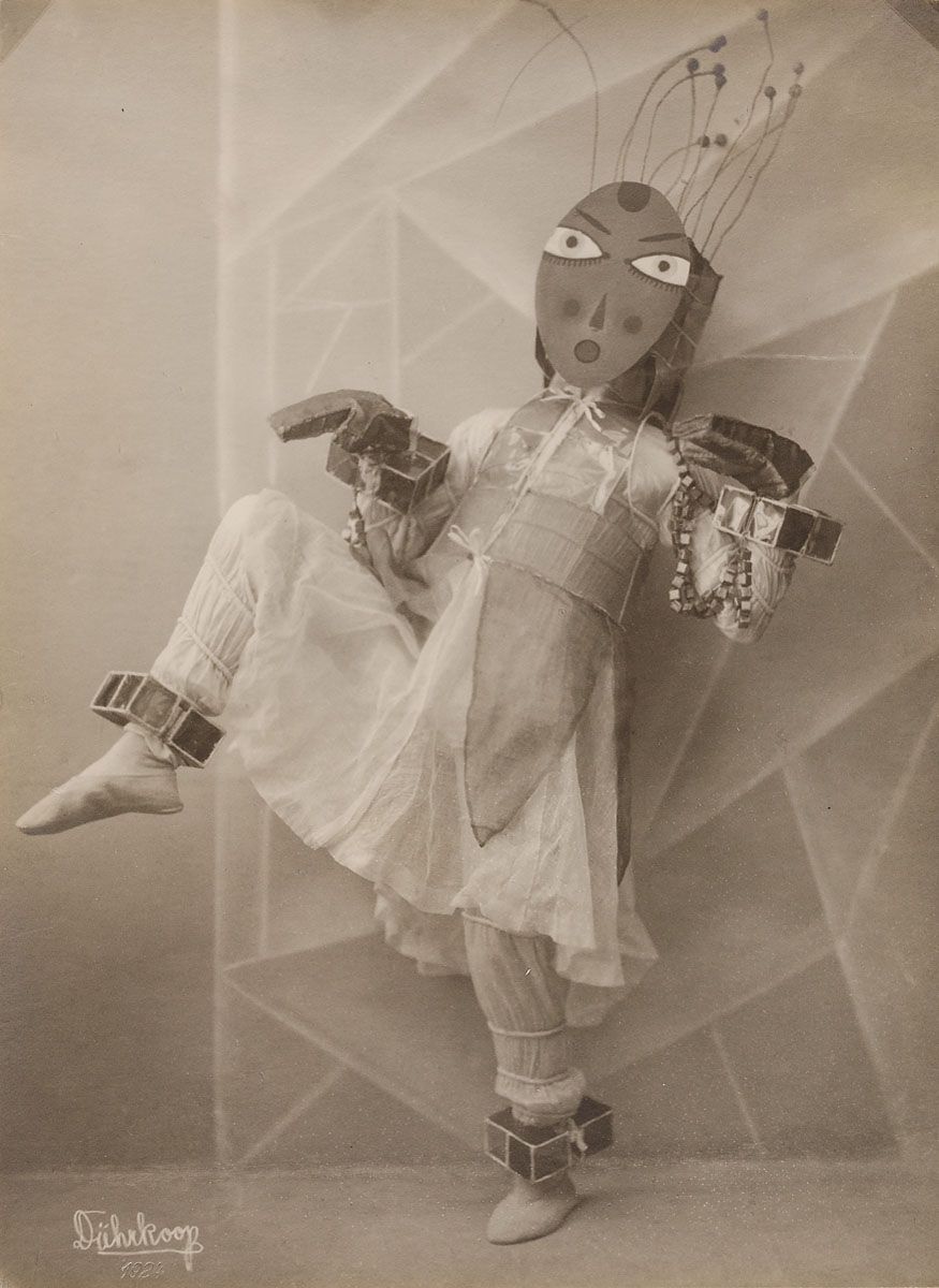 A sepia-toned photograph of a figure in a surreal costume, featuring a rectangular mask with wire-like extensions, a ruffled white garment, and numerous box-like structures attached to the costume. The figure is posed dynamically against a geometric background. Signed 'Dahukoog' 1922.