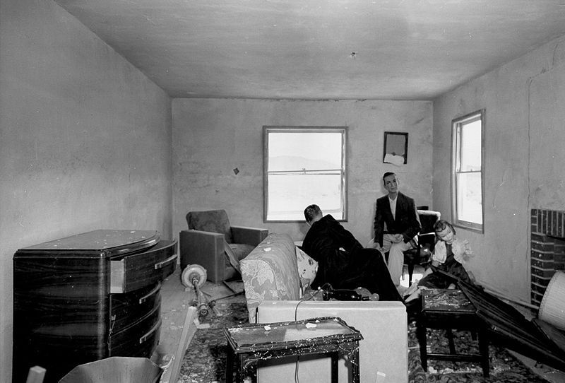 A black and white photograph of a ransacked living room with two men surveying the damage.