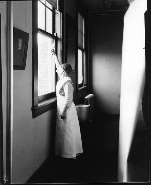 A black and white photograph of a woman in a long dress reaching up to adjust a window in a simple room. A beam of light streams in, creating a dramatic effect.