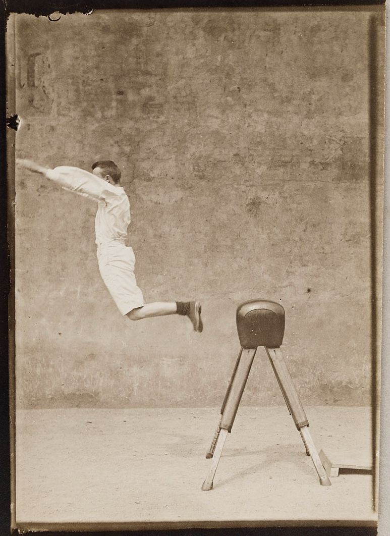 Vintage photograph of a young man performing a gymnastic vault over a wooden horse in a gymnasium.