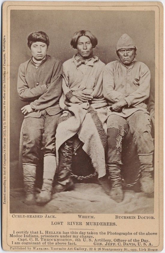 A sepia-toned photograph of three Native American men labeled as “Lost River Murderers.” They are standing in a studio setting, wearing a mix of traditional and Western clothing. The photograph is a historical document with text at the bottom certifying the authenticity of the image.