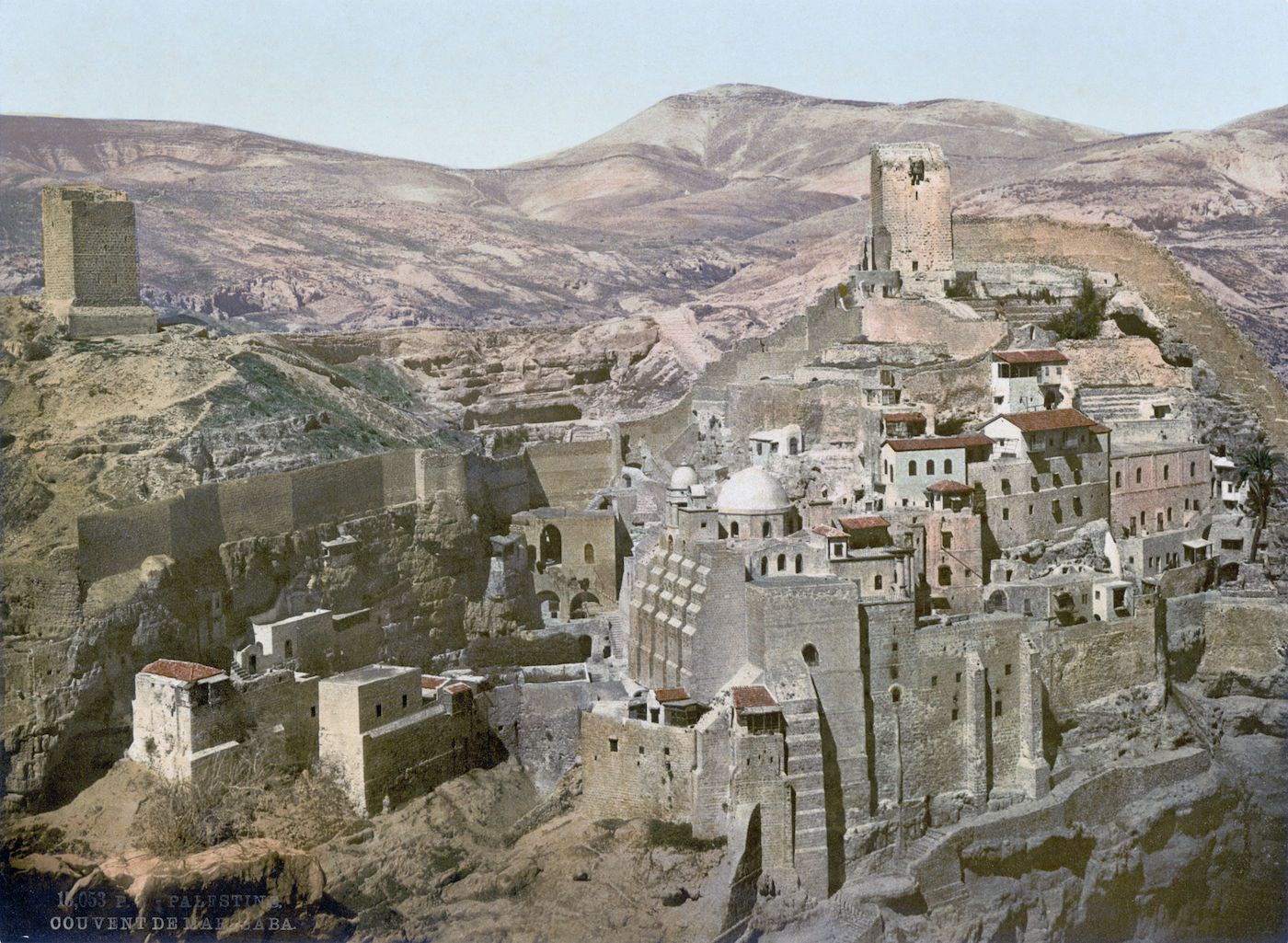 Vintage colorized photograph of the Mar Saba Monastery in Palestine, a stone structure built into a rocky hillside with towers and surrounded by a barren landscape.