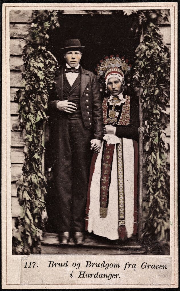 Vintage photograph of a bride and groom in traditional Norwegian folk costumes, standing in front of a wooden building covered in greenery.