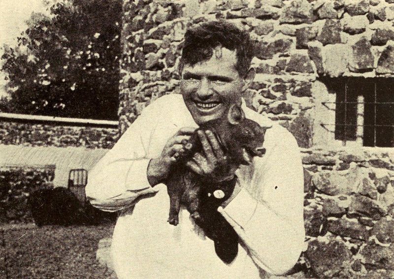 A black and white photograph of a smiling man holding a small dog in front of a stone wall.