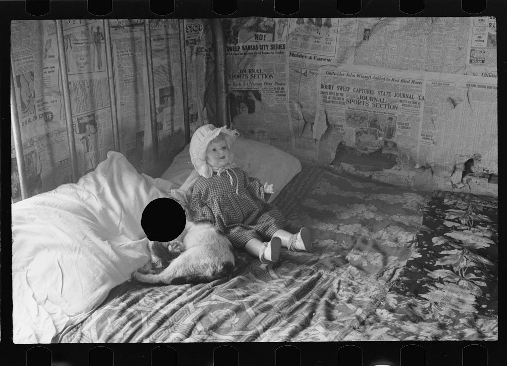A black and white photograph of a young girl sitting on a bed in a room with walls covered in newspapers.