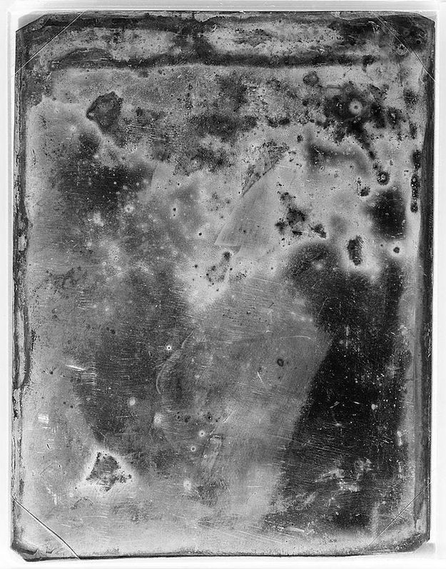 A heavily tarnished and damaged daguerreotype, showing a vague suggestion of a human face or figure amidst a swirling mass of grays and blacks.