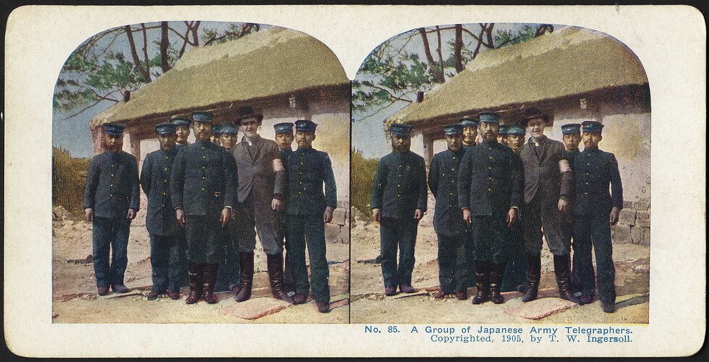 Stereoscopic photograph of a group of Japanese Army Telegraphers in uniform, standing in front of a wooden structure, 1905.