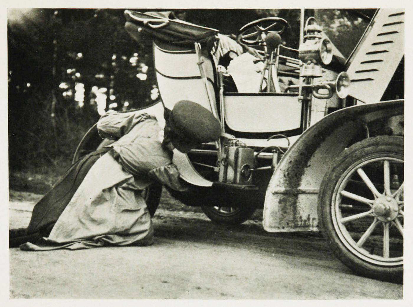 A vintage black and white photograph of a person working on the front tire of a classic car on a dirt road.