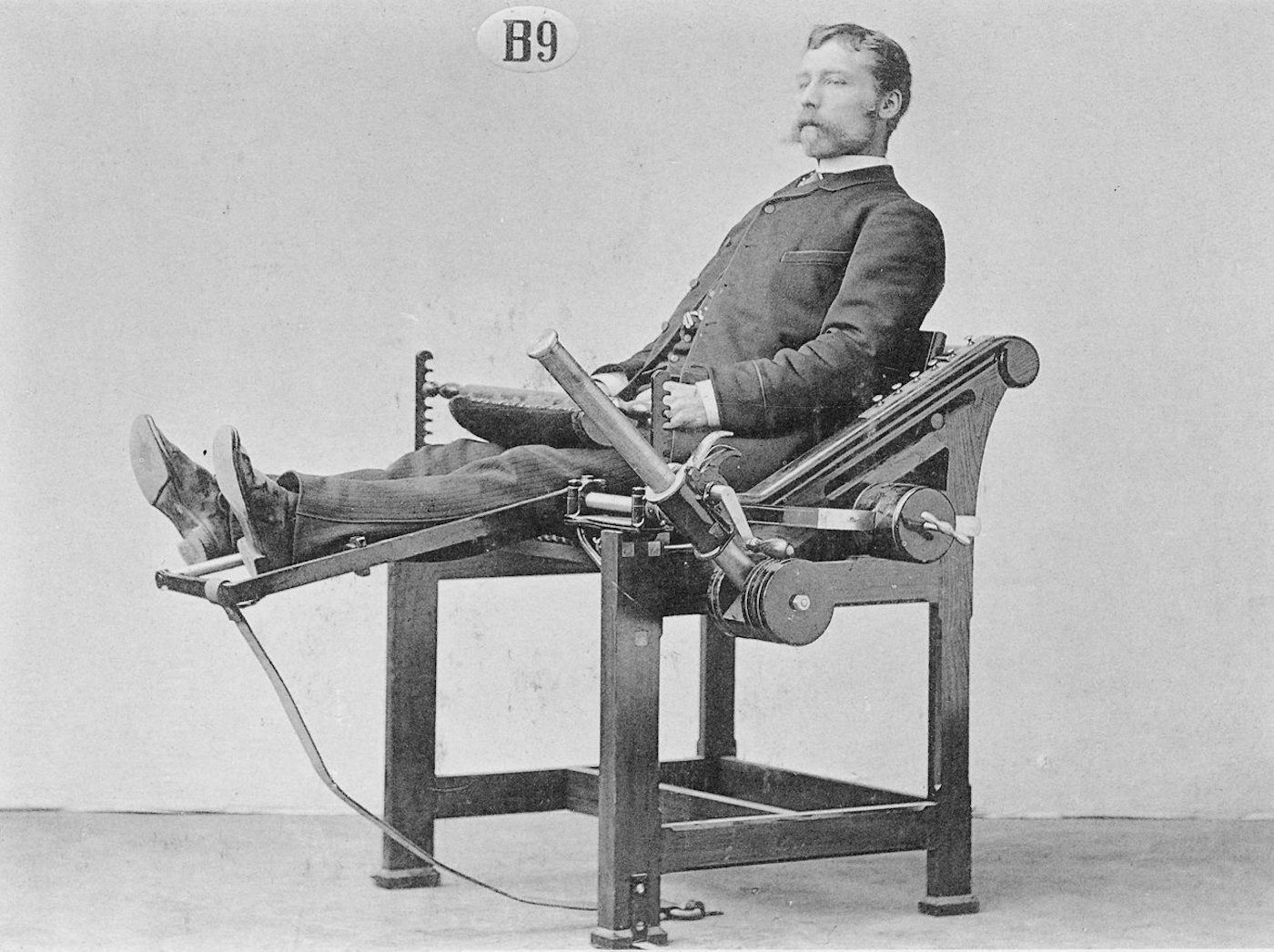 A black and white photograph of a man seated in a complex mechanical chair, likely a medical or therapeutic device from the late 19th or early 20th century.