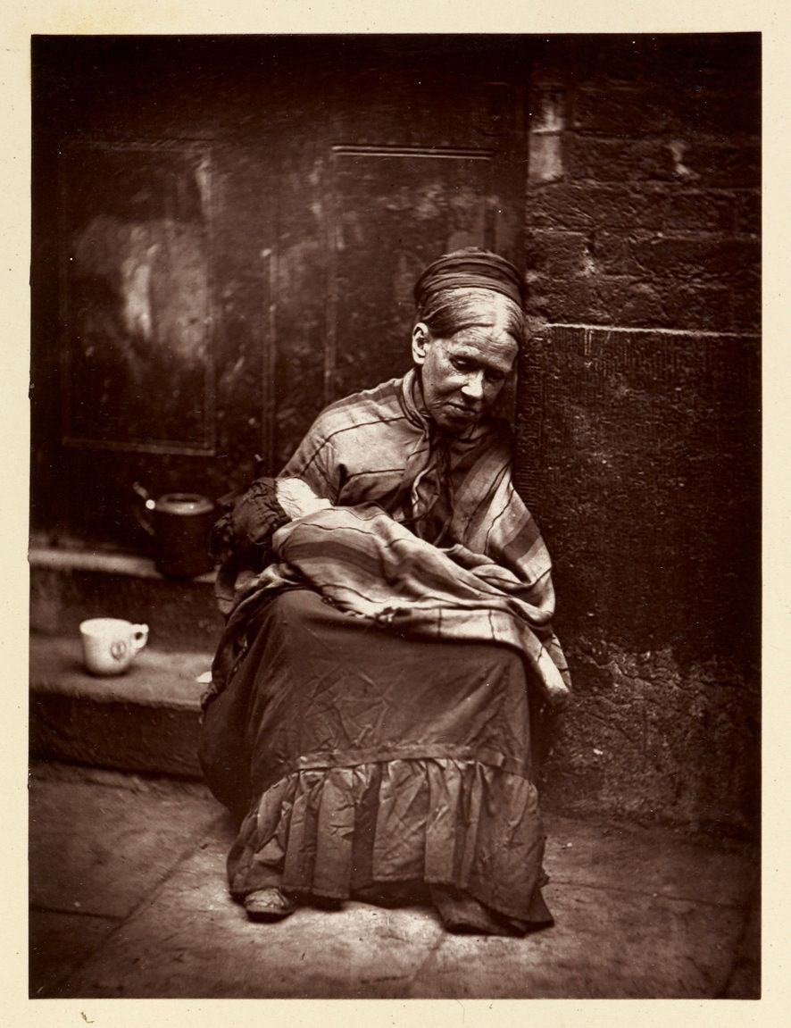 A vintage black and white photograph of an elderly woman seated indoors, looking down with a somber expression. She is wearing a head covering and a long dress, clutching something in her hands. A cup and pot sit on a surface to her left, and a textured wall forms the background.