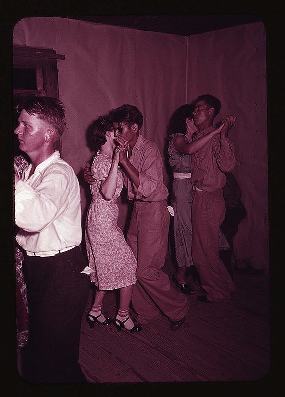 A vintage photograph of three couples dancing closely in a dimly lit room. The couples are dressed in mid-20th-century attire, and the room is decorated with a simple pinkish-red fabric. A man stands to the left, observing the scene.