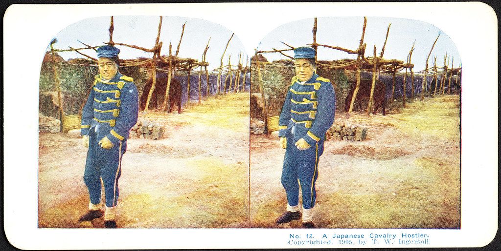 Stereoscope card of a Japanese cavalry hostler in uniform standing in front of thatched huts.