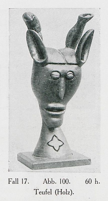 A black and white photograph of a wooden sculpture of a devil with curved horns and a stylized face.