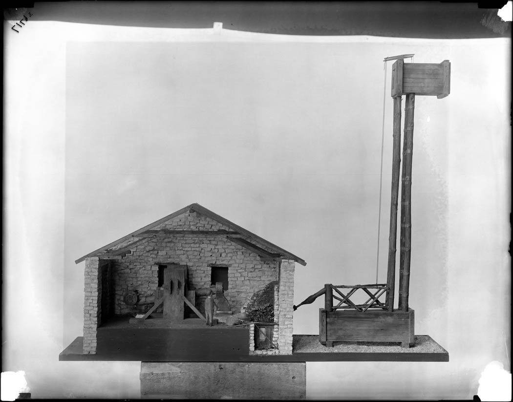 Black and white photograph of a miniature model of a stone building and a tall wooden tower on a flat base.