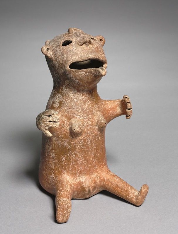 A terracotta figurine of a creature with a humanoid form and animalistic features, seated with its legs bent and arms raised.