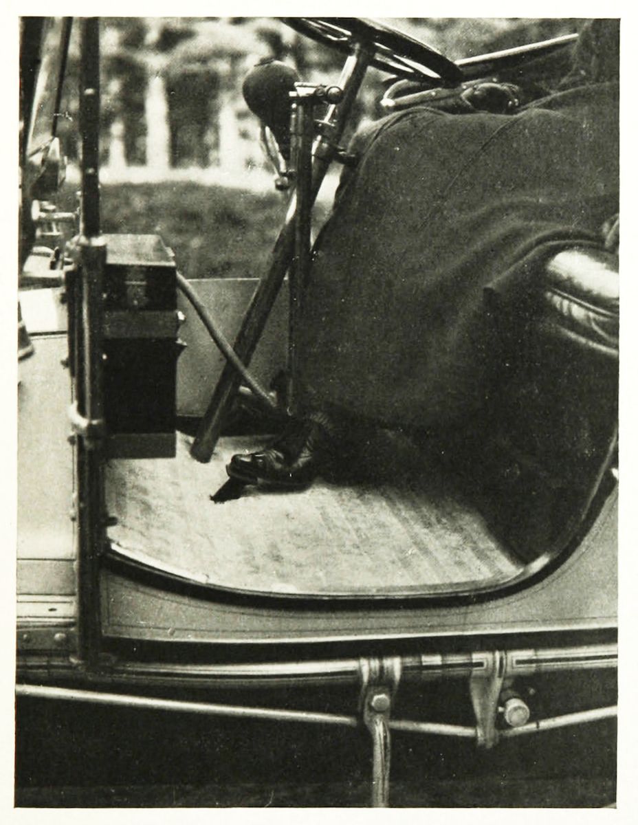 Vintage black and white photograph of a classic car interior, showing the steering wheel, seat, and pedals, with a blurred view of trees and a building through the window.