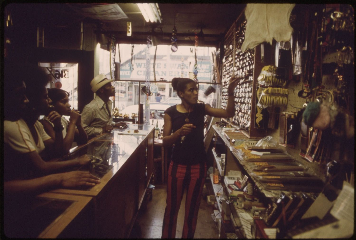 Interior of a jewelry store with a long counter, several customers, and a shopkeeper gesturing with his hand.