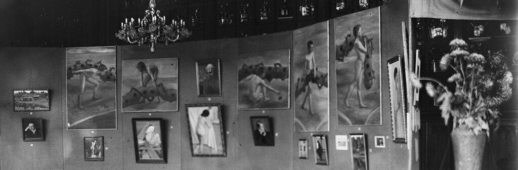 A black and white photograph of an art gallery interior with numerous paintings on the walls and a chandelier hanging from the ceiling.