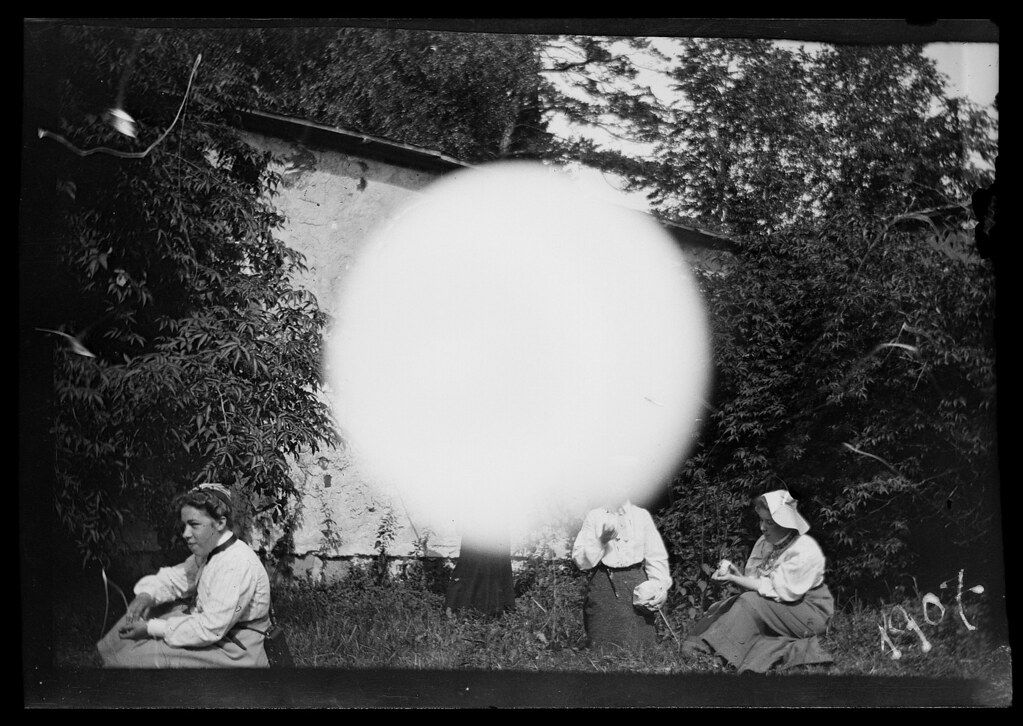 A black and white photograph of three people seated in a grassy area in front of a building, with a large, bright circular shape dominating the center of the image.