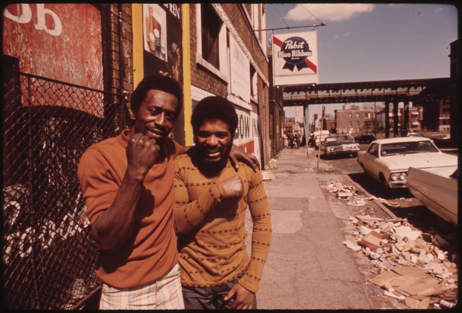 Two young African American men stand smiling in front of a brick building in a rundown urban neighborhood.