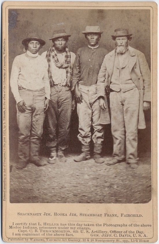 A vintage photograph of four Native American men standing in a row, posing for the camera. They are dressed in a mix of traditional and Western clothing. The photograph is sepia-toned and has a slightly grainy texture. There is text at the bottom of the image that identifies the men as Shacknasty Jim, Hooka Jim, Steamboat Frank, and Fairchild. It also includes a certification from Capt. C. B. Throckmorton and Gen. Jeff. C. Davis, U.S.A., stating that the photograph was taken of Modoc Indians who were prisoners under their charge. The photograph was published by Watkins, Yosemite Art Gallery.