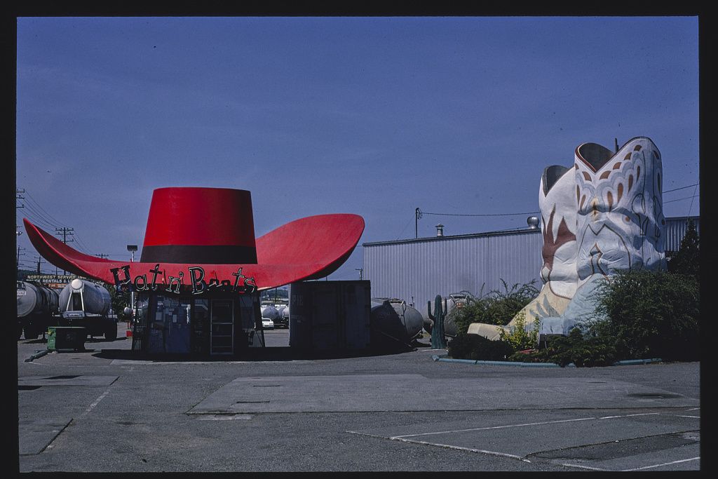 A vintage roadside attraction featuring a large red cowboy hat and a giant sculpted cowboy boot, set in an empty parking lot under a clear blue sky.